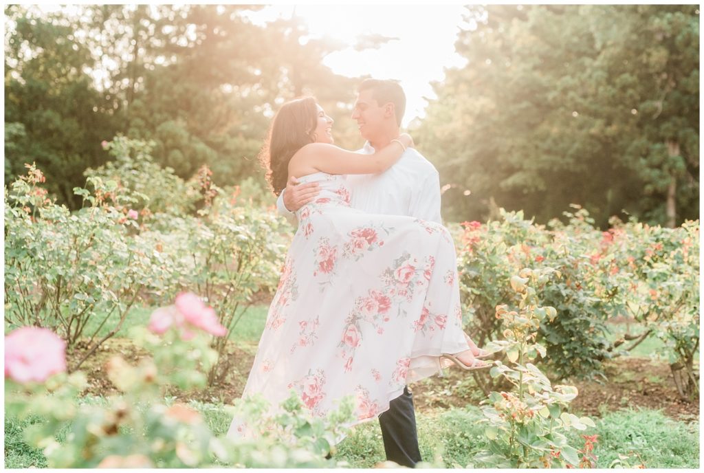 A man carries his fiance through a rose garden in glowy golden light.