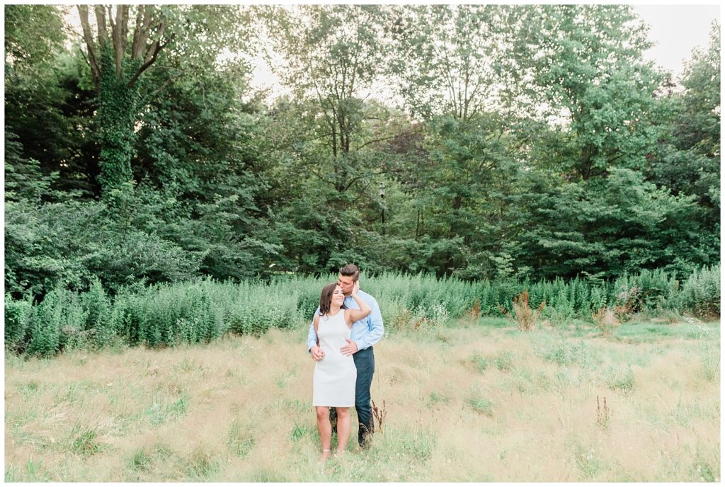 A man hugs his fiancee from behind in a field of tall grass.