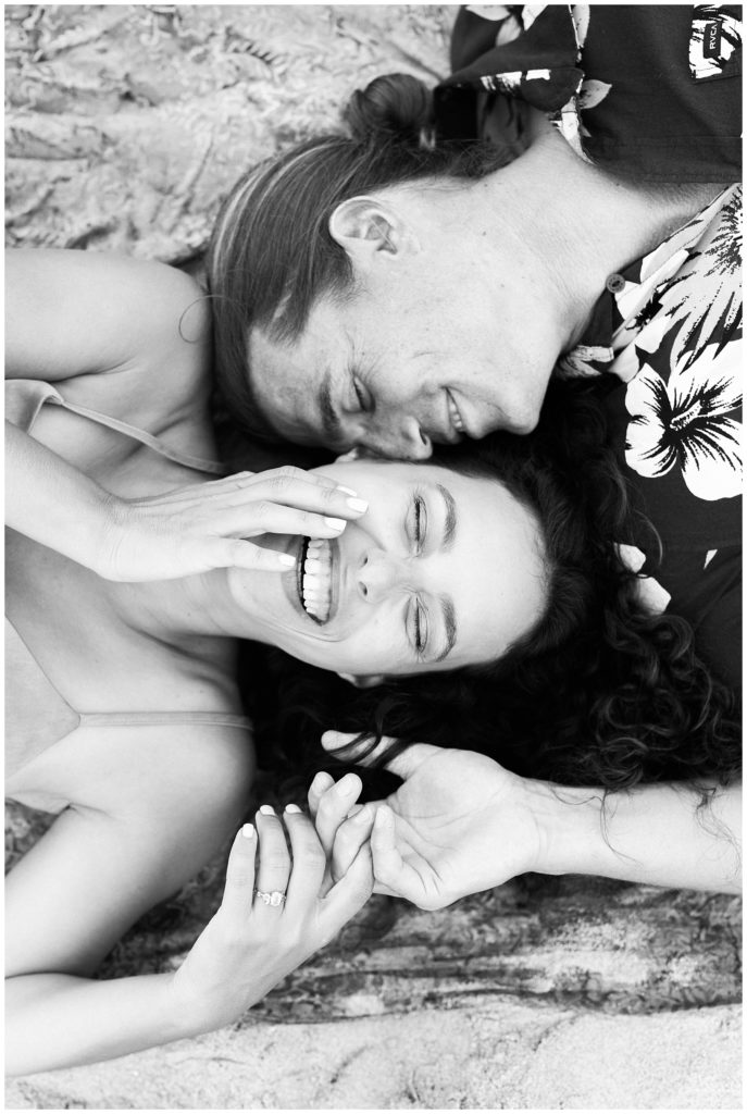 A black and white image of Thiana and Brandon laying on their backs laughing. He is looking at her and she is facing straight up and has her right hand resting on her face. 