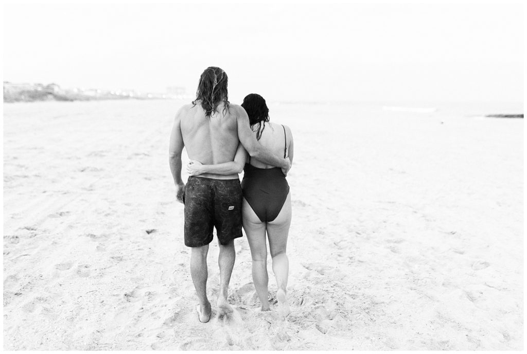 A black and white image of Thiana and brandon. They are walking away from the camera with their arms around each other along the shoreline. Thiana's sholders are slightly hunched. 
