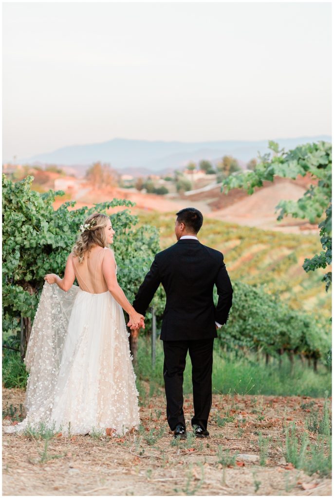 A bride and groom hold hands overlooking the rolling hills of the vineyard at Avensole Winery wedding venue in Temecula, CA.