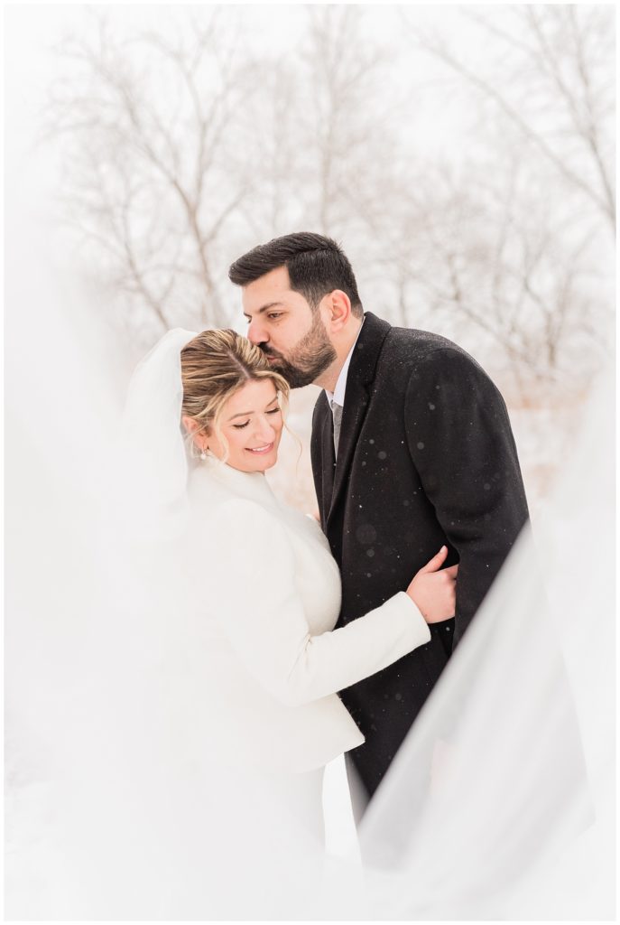 A groom kisses the bride on her temple with her veil sweeping in front as the snow falls around them in Beacon NY at Roundhouse Hotel winter wedding.