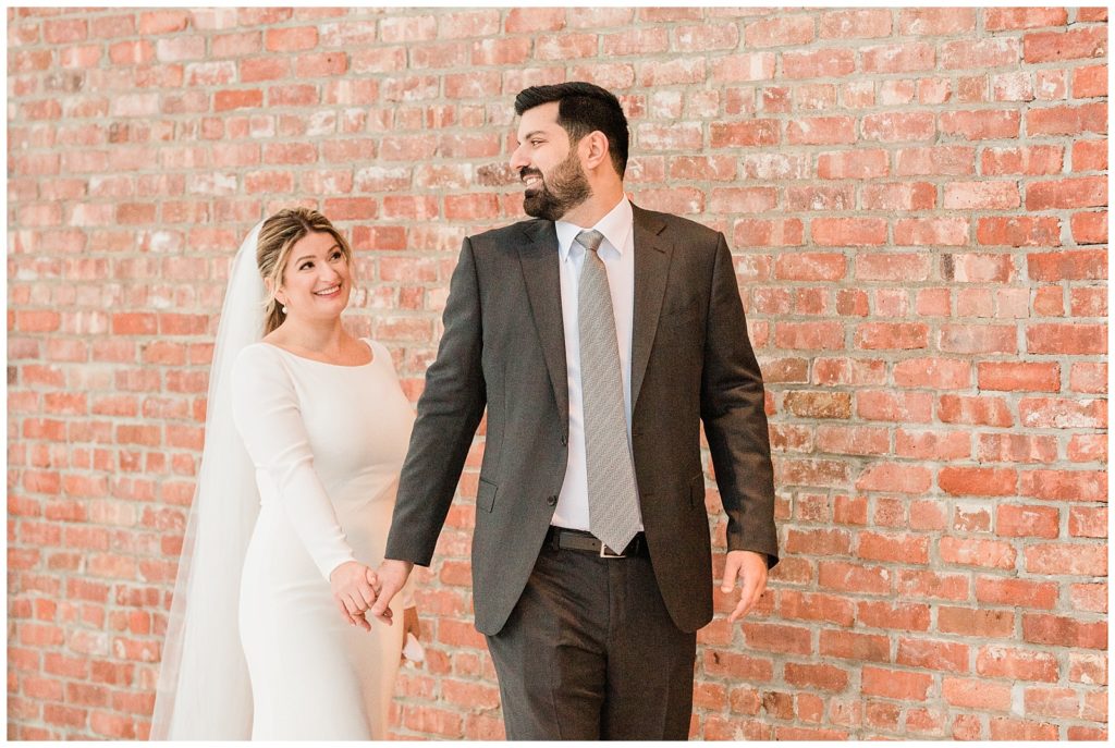 A groom holds his bride's hand and walks in front of a brick wall in Beacon NY at Roundhouse Hotel winter wedding inspiration.