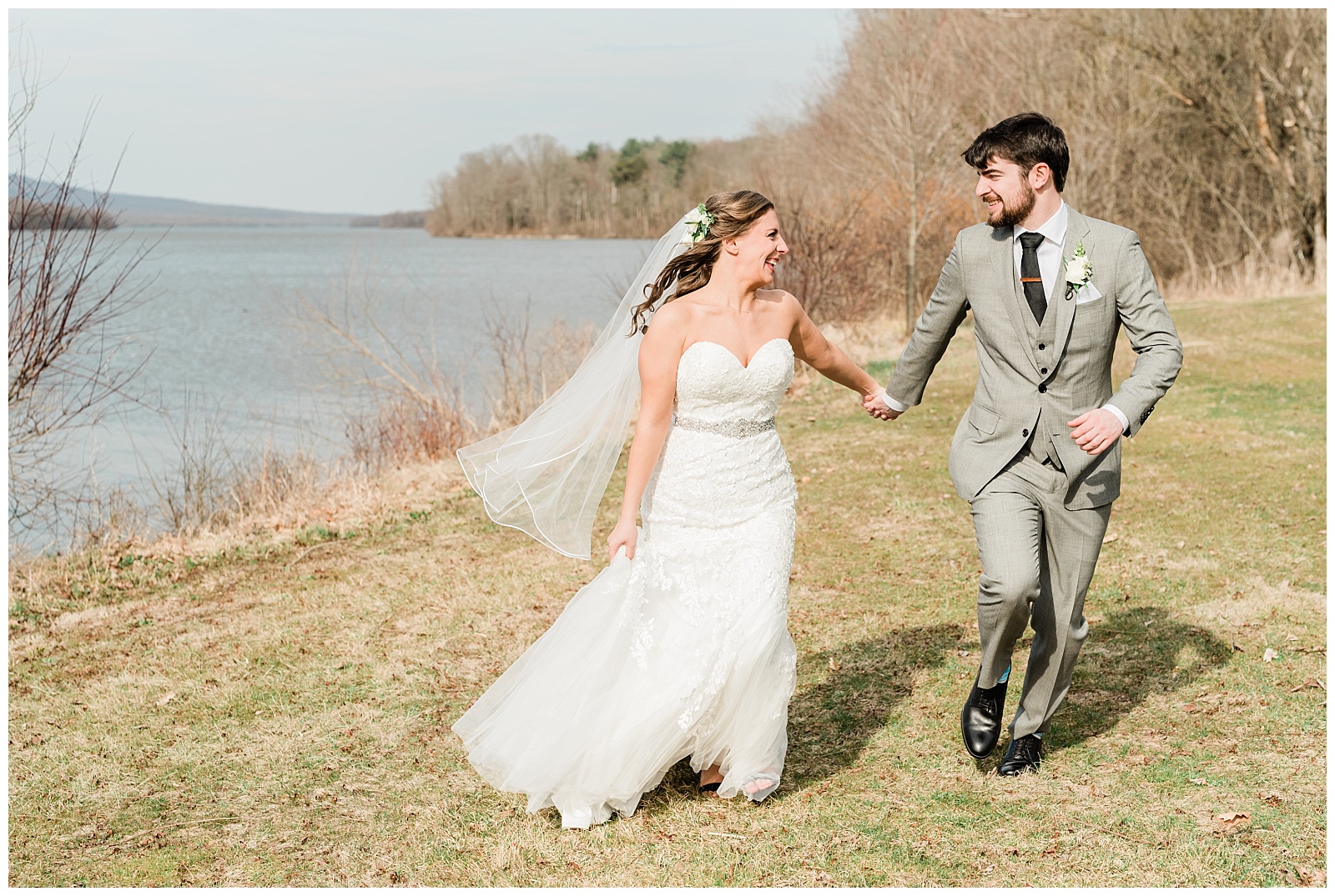 Bride and groom holding hands running next to a lake.