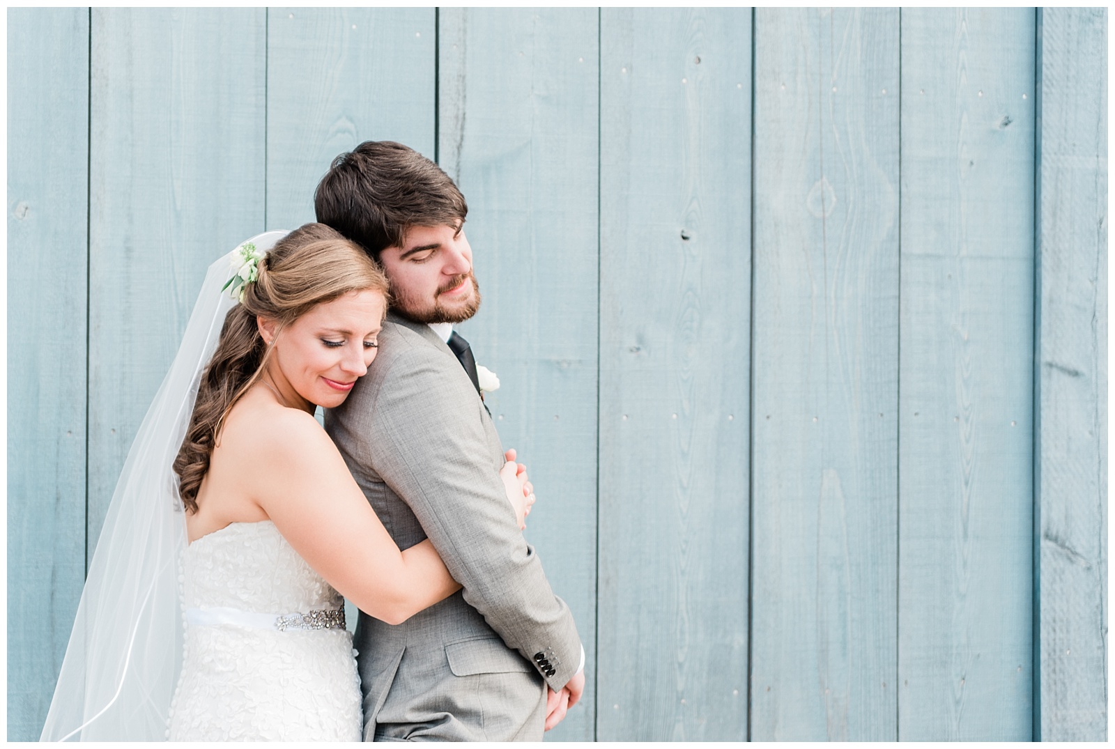 Bride hugging her groom from behind and resting her head on his back in front of a blue wall.