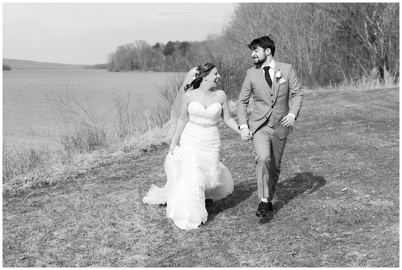 Black and white portrait of a bride and groom running next to a lake in a Mark Zunino gown.