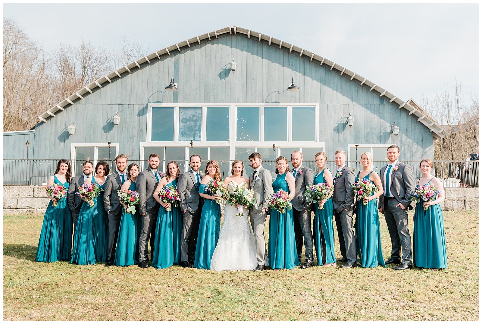 A wedding party of bridesmaids wearing teal dresses and groomsmen wearing grey suits, in front of the Lake House Inn wedding venue in Pennsylvania.