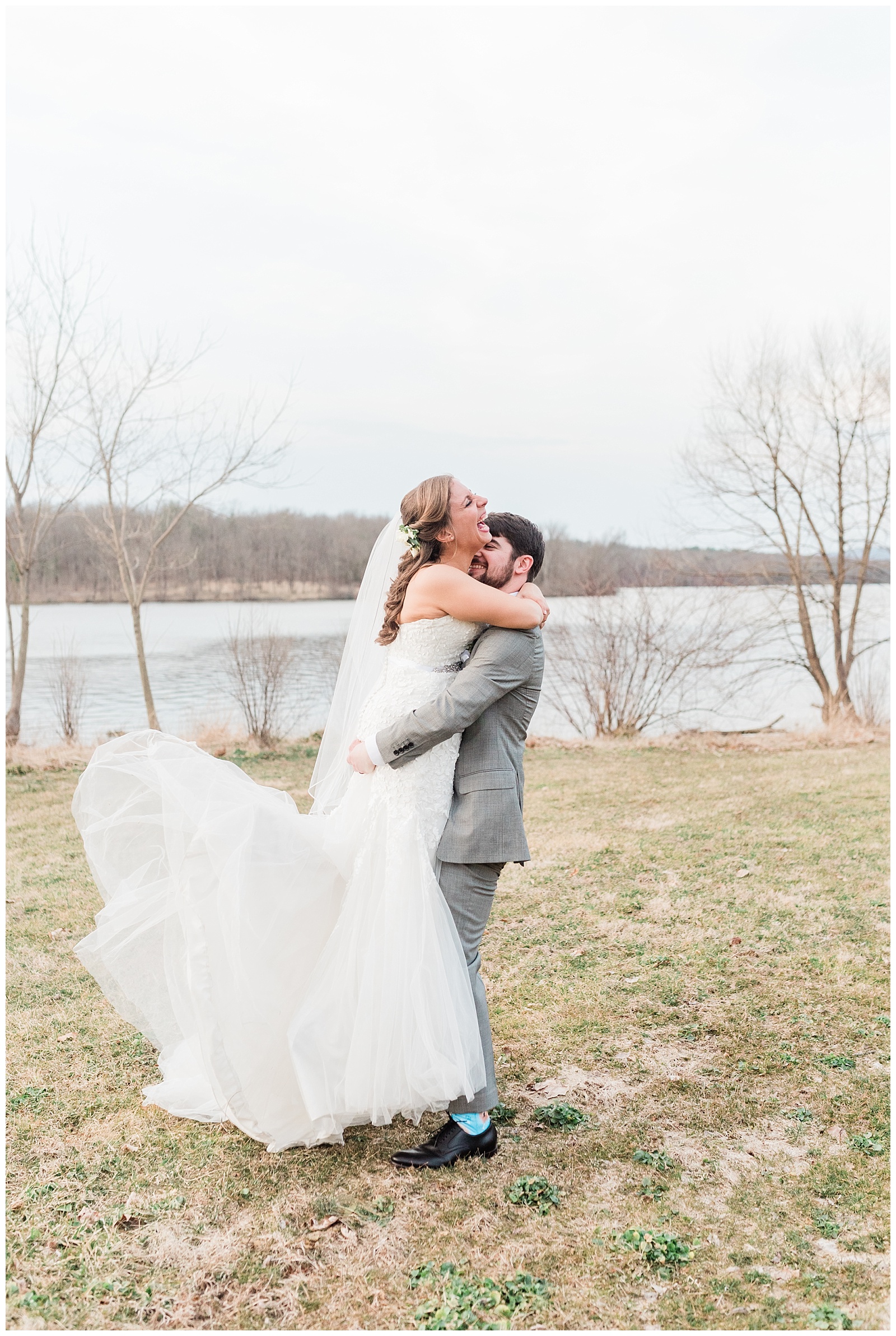 Bride laughs as the groom lifts her up in front of a lake, and she kicks up her Mark Zunino wedding gown.