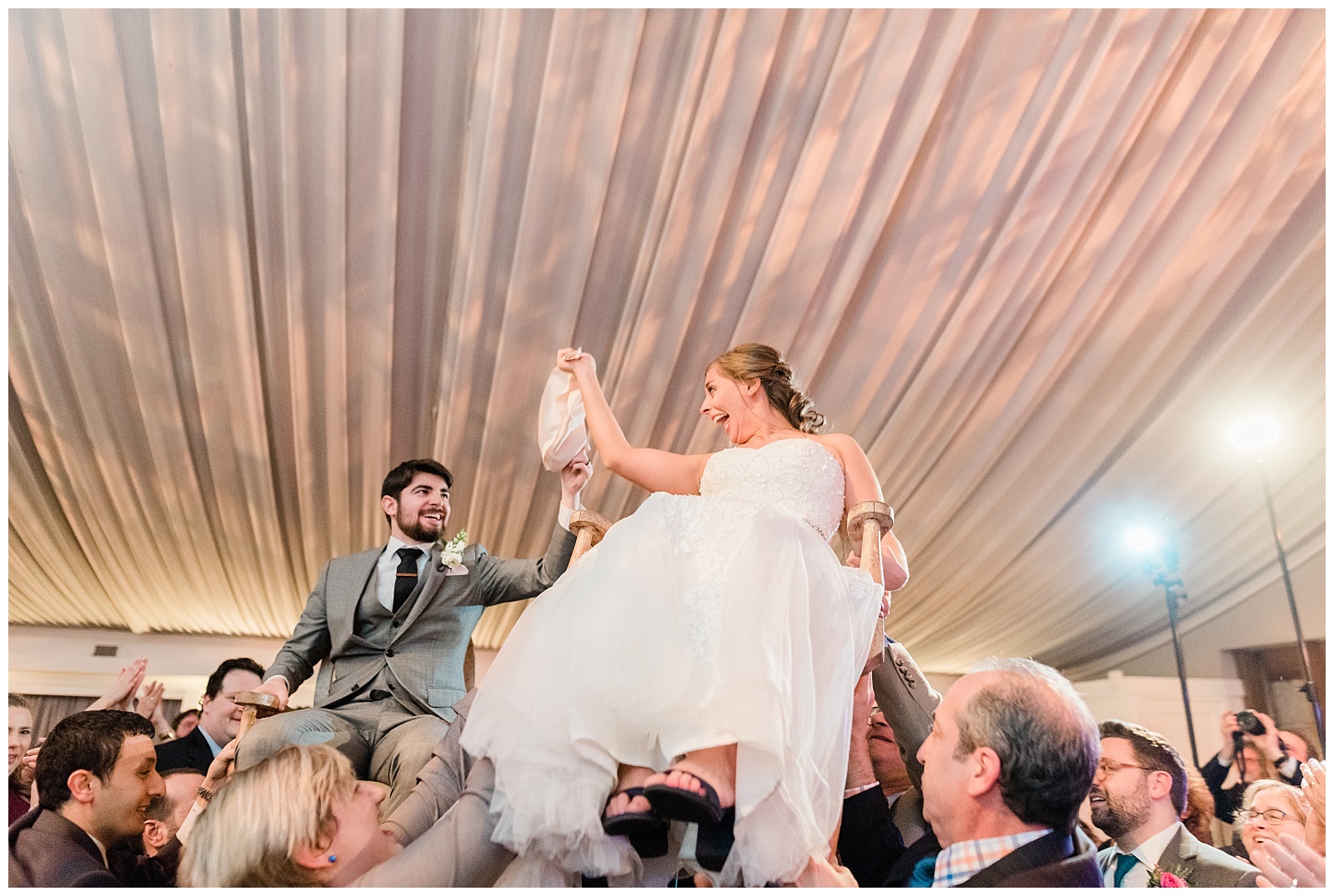 Bride and groom hold a napkin while being lifted on chairs for the Jewish hora dance at their wedding.