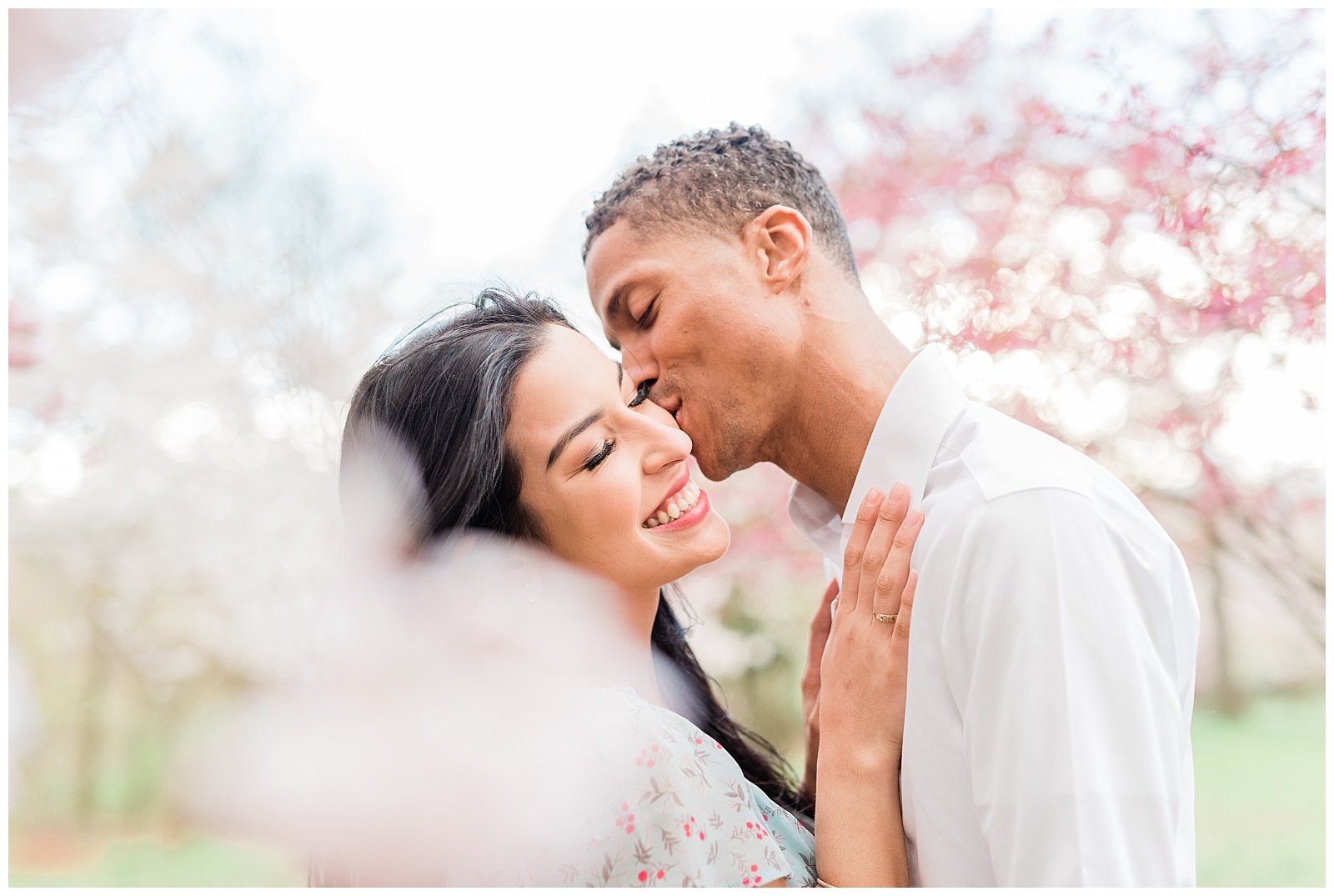 A man kisses his fiance on the cheek among the cherry blossom trees.