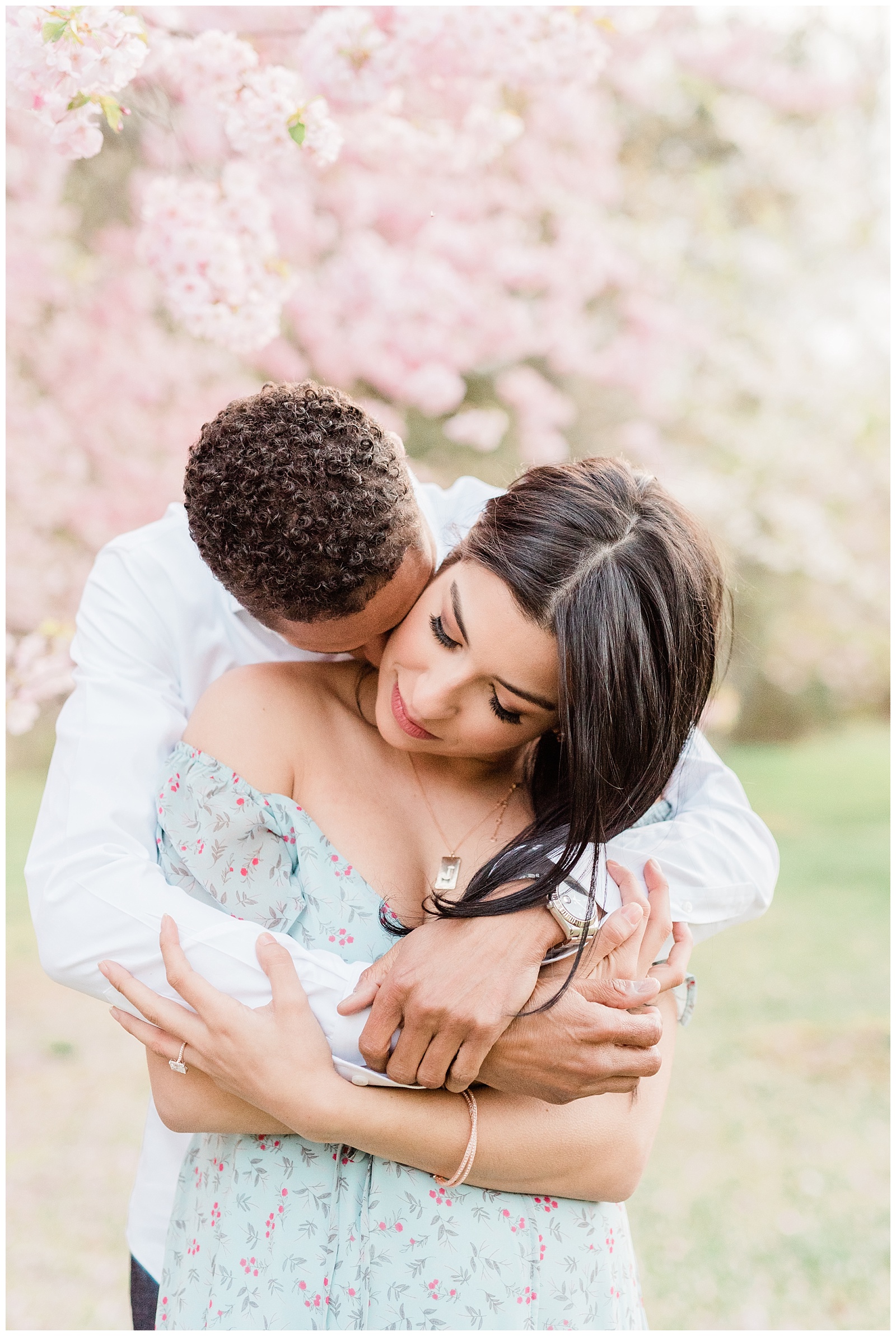 A man hugs his fiance from behind in a cherry blossom field at springtime.