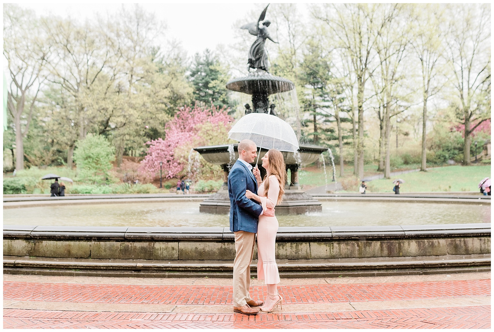 A couple smiles at each other while sharing an umbrella in front of Bethesda Fountain in Central Park.