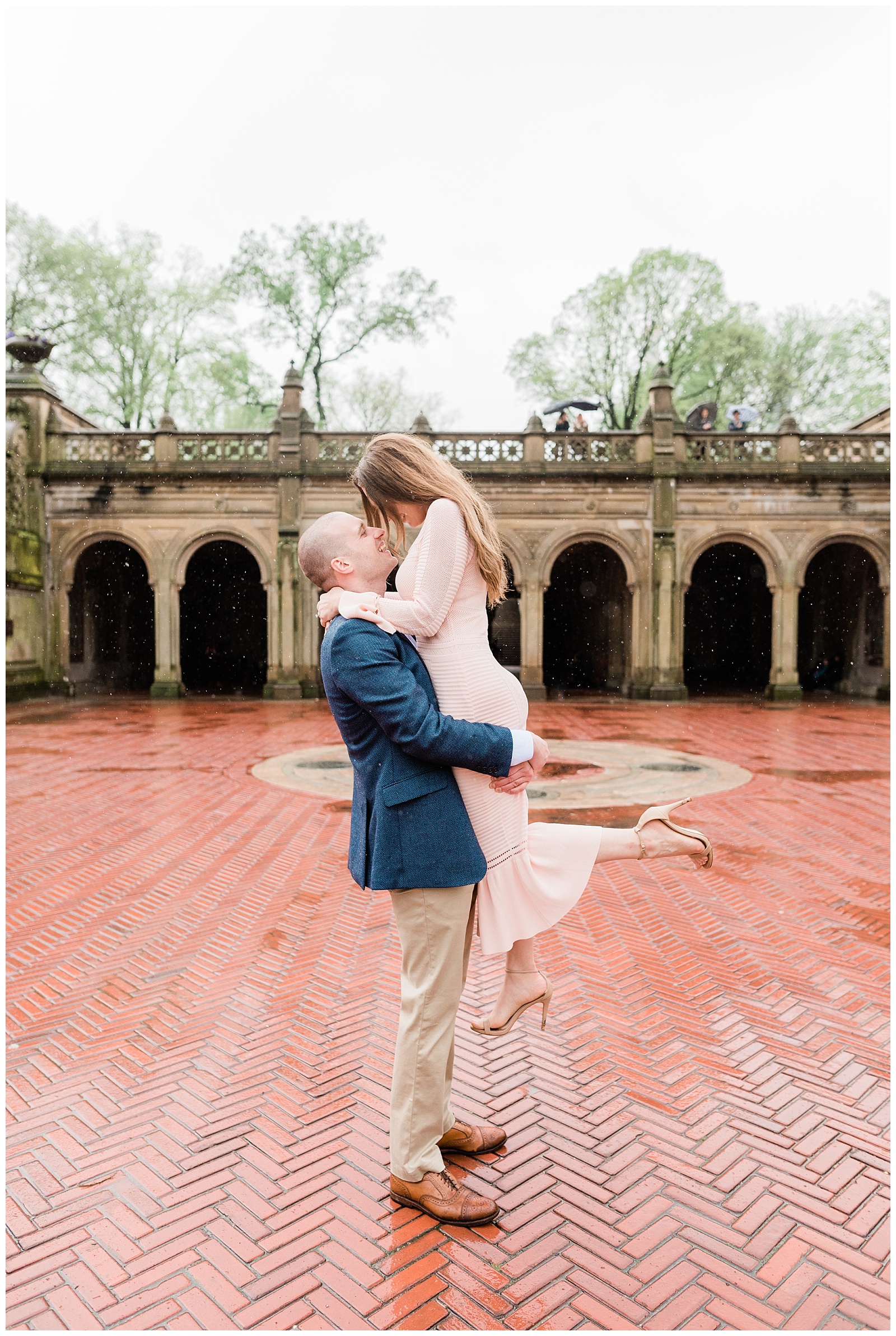 A man lifts up his fiancee in front of Bethesda Terrace in the rain.