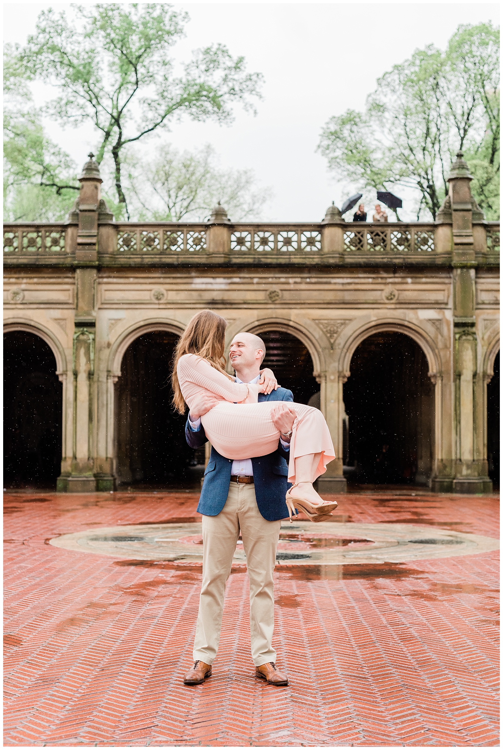 A man holds a woman in front of Bethesda Terrace in the rain.