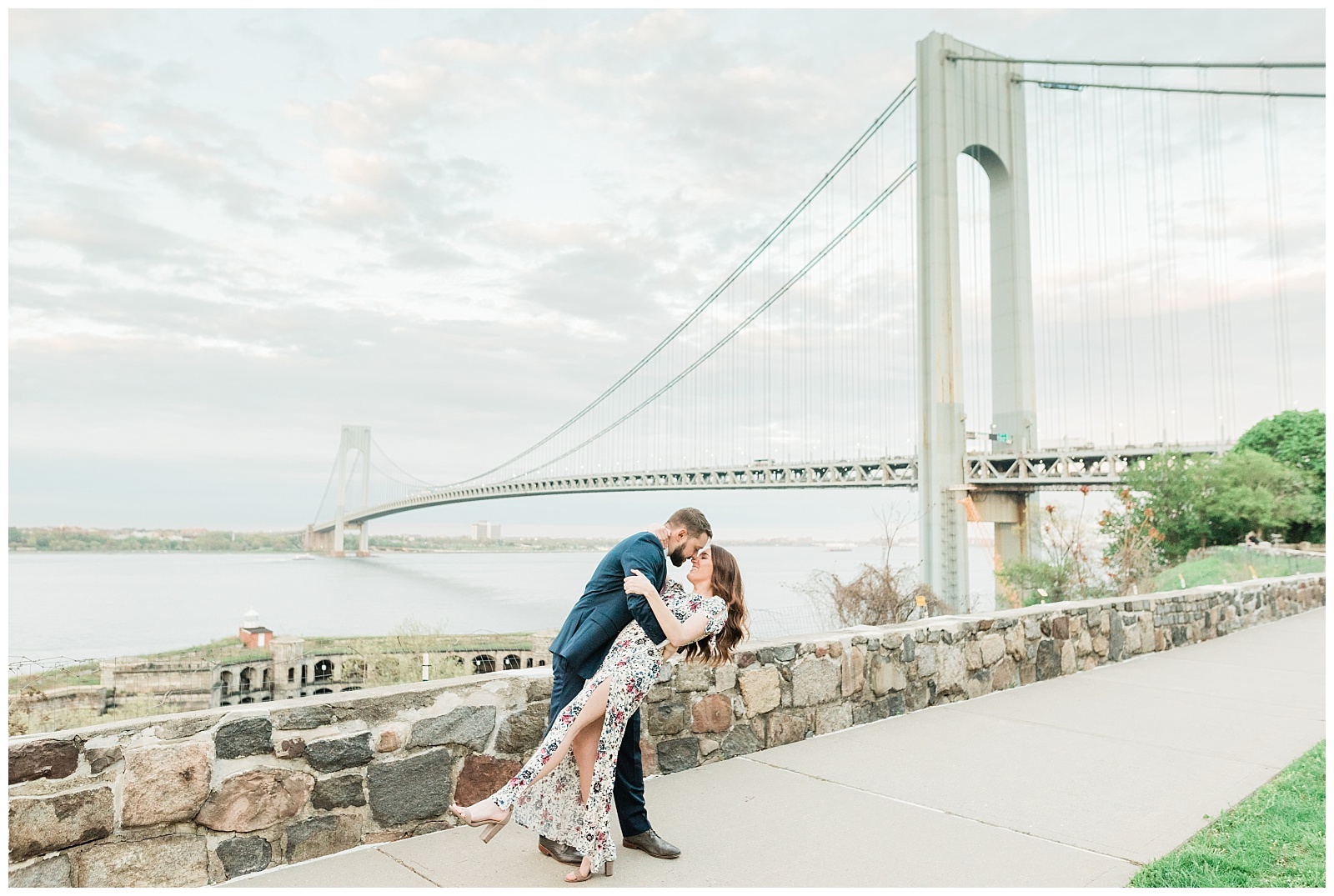 A man dips a woman back and kisses her in front of the Verrazzano Bridge.