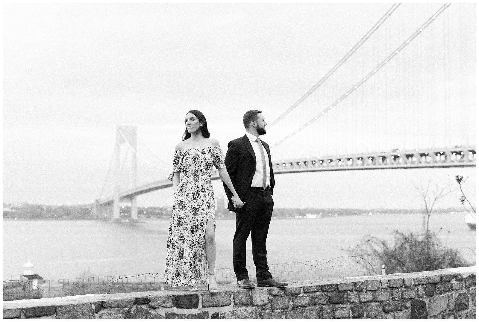 A couple holds hands, and stands along the ledge of a stone wall by the Verrazzano Bridge.