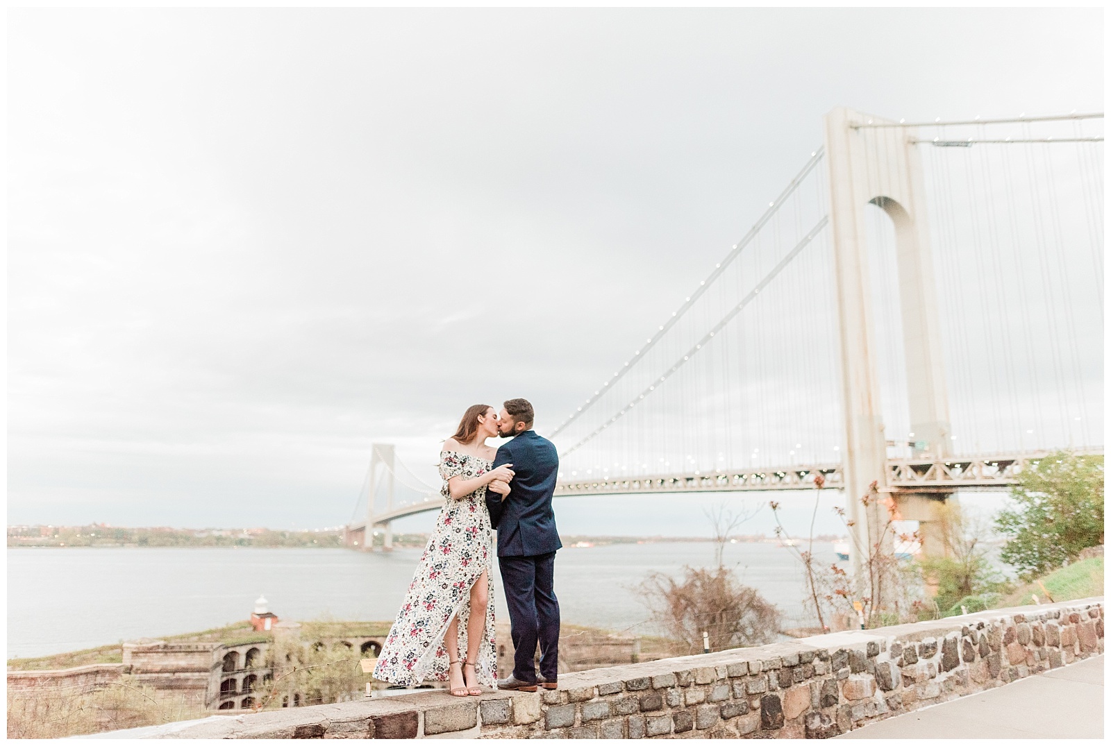 A couple kisses on a stone ledge by the Verrazzano Bridge.