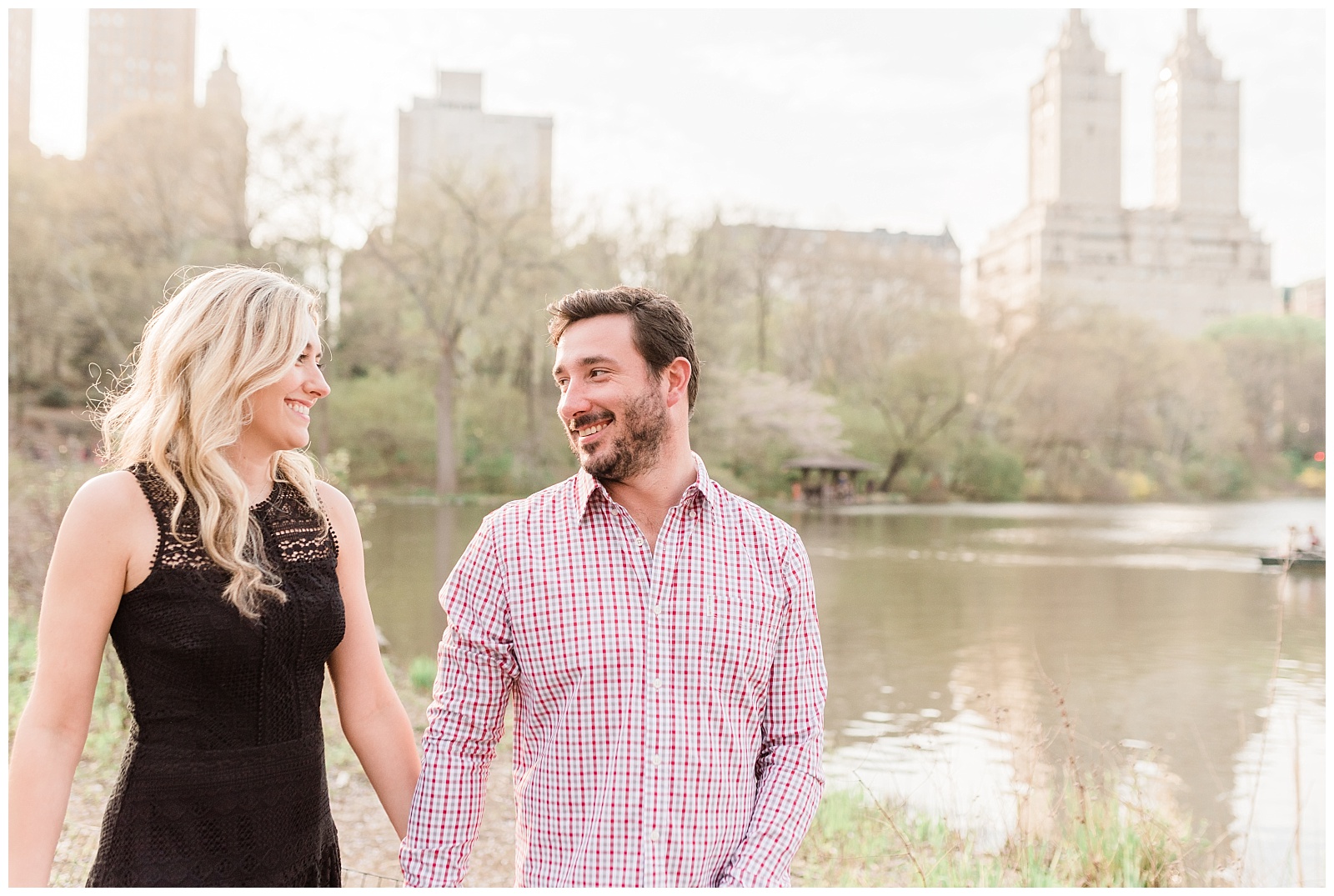 A couple smiles at each other, walking in front of the lake in Central Park.