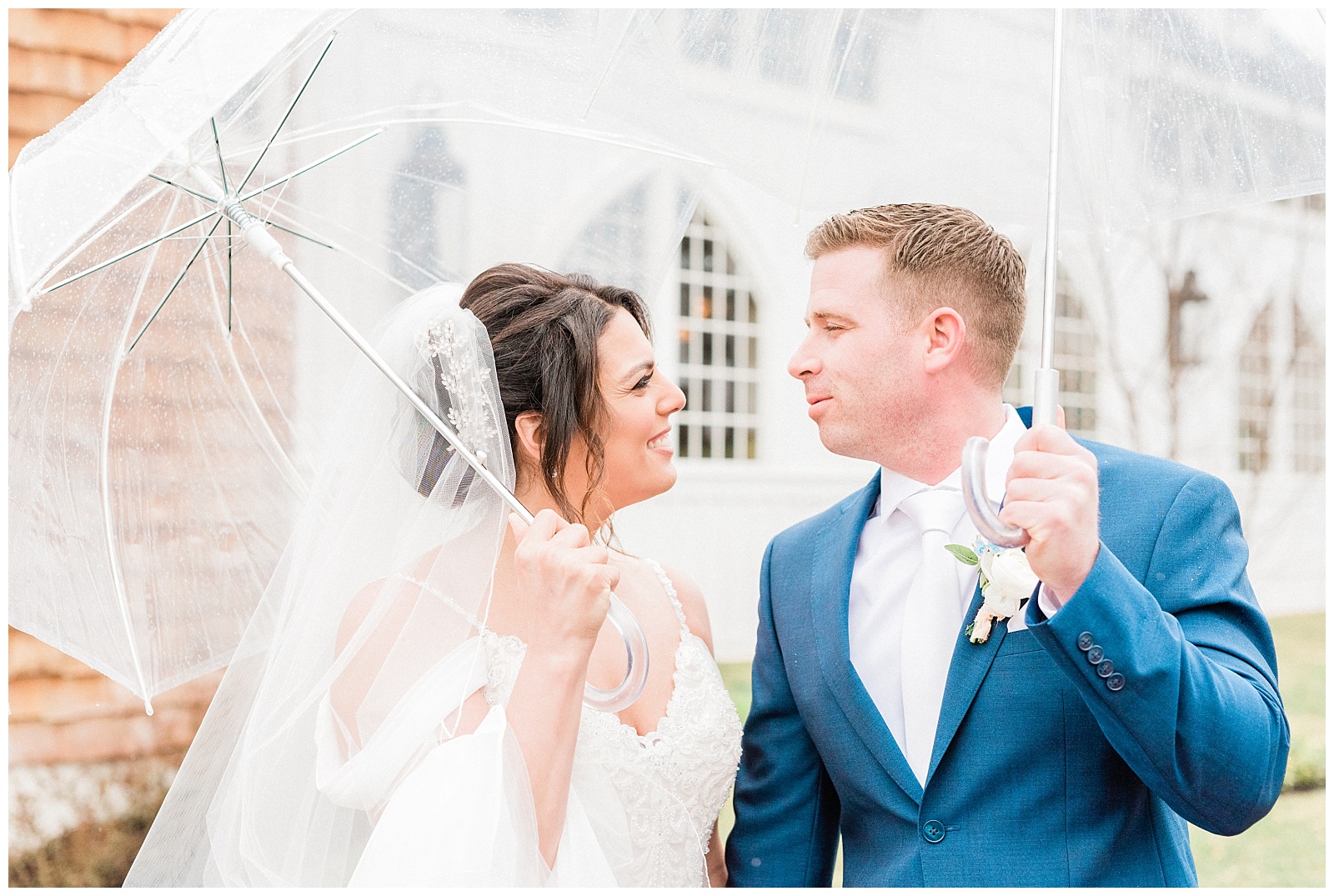 Bride and groom look at each other while holding clear umbrellas in the rain.