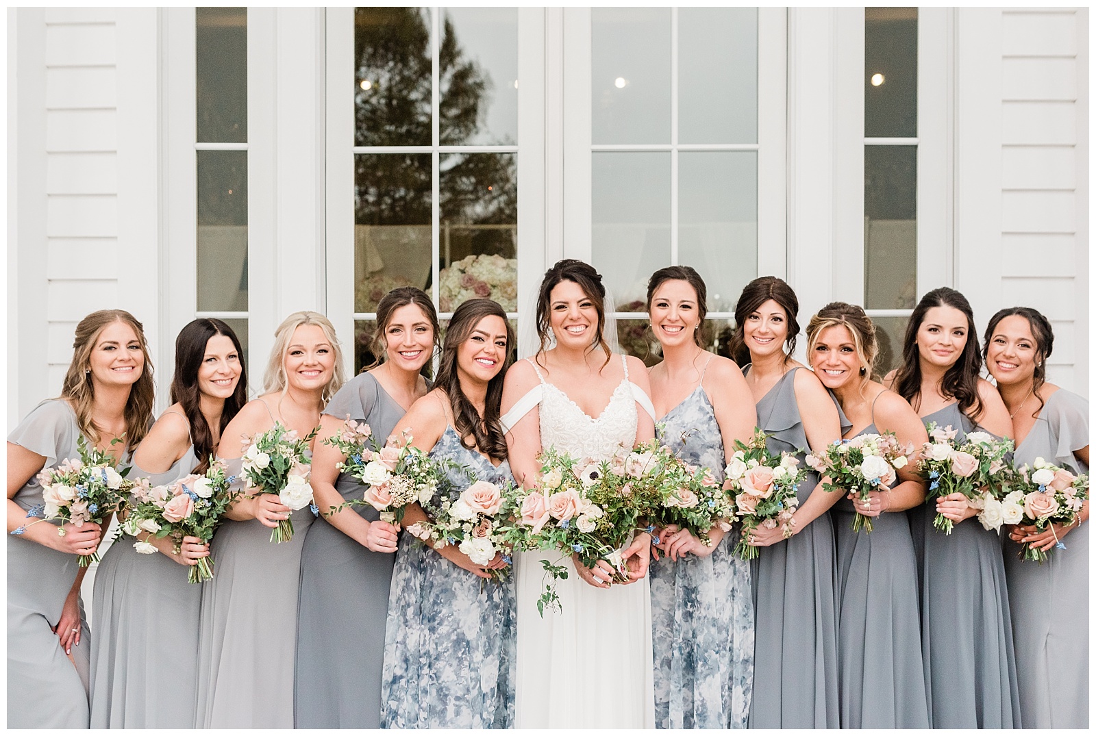 Bridesmaids wearing different shades of grey dresses hold their bouquets and smile.