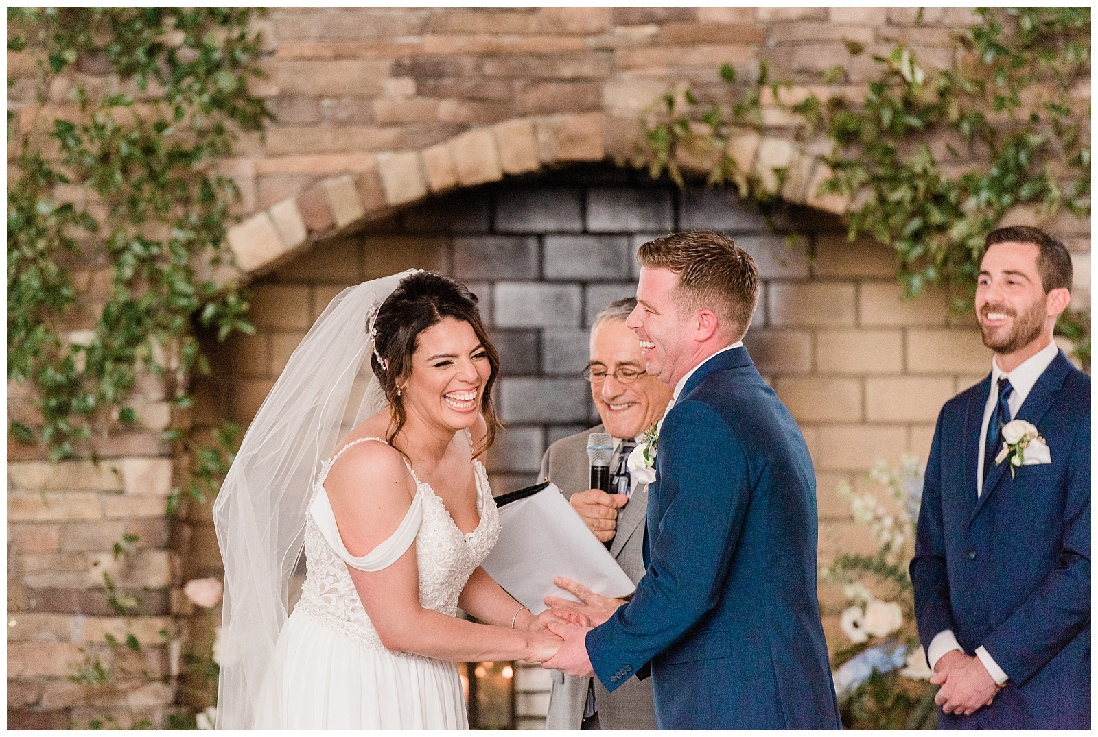 Bride and groom laugh while holding hands during the ceremony in front of a fireplace.