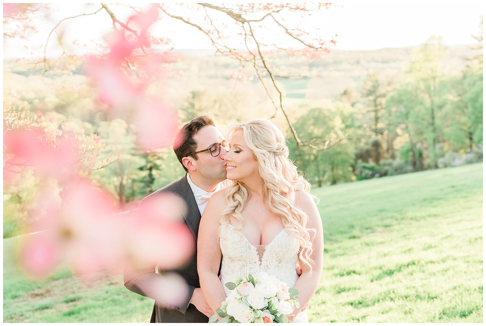 A groom kisses his bride on the cheek at golden hour underneath a pink dogwood tree.