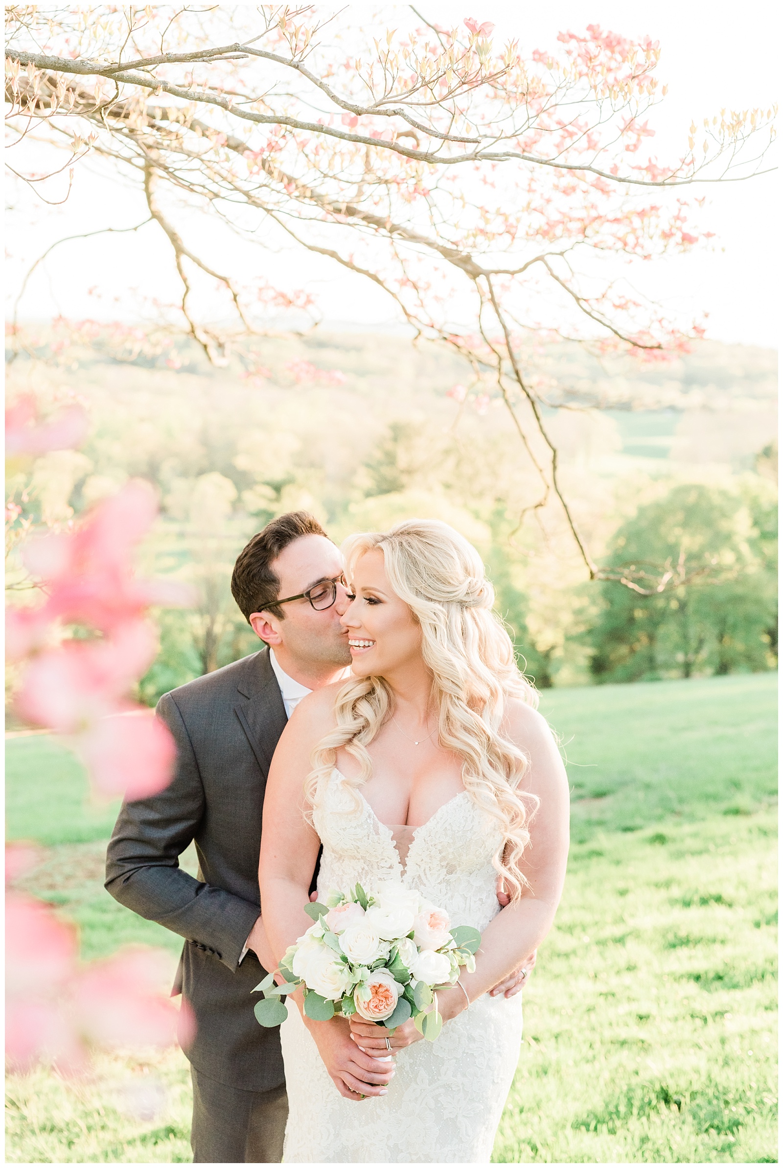A groom kisses his bride on the cheek at golden hour underneath a pink dogwood tree.