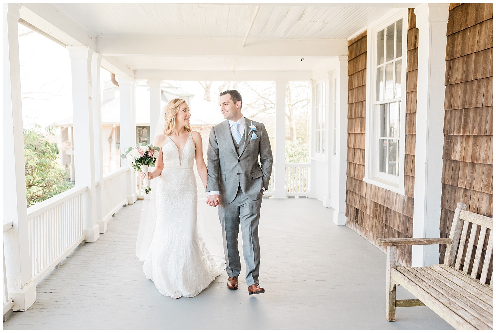 Bride and groom walk holding hands on a covered porch.