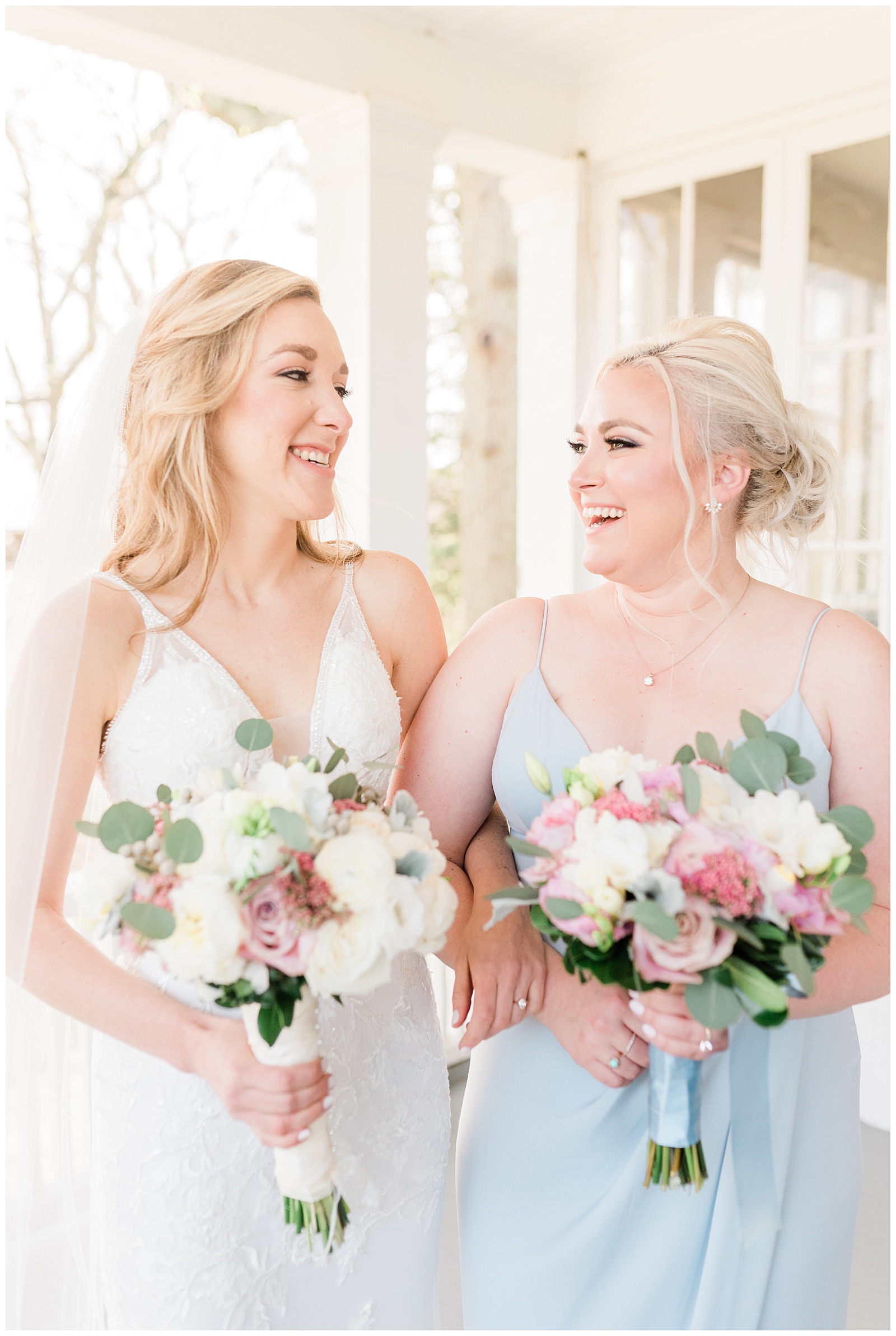 Bride and maid of honor laugh while holding their bouquets of white and pink flowers and eucalyptus leaves.