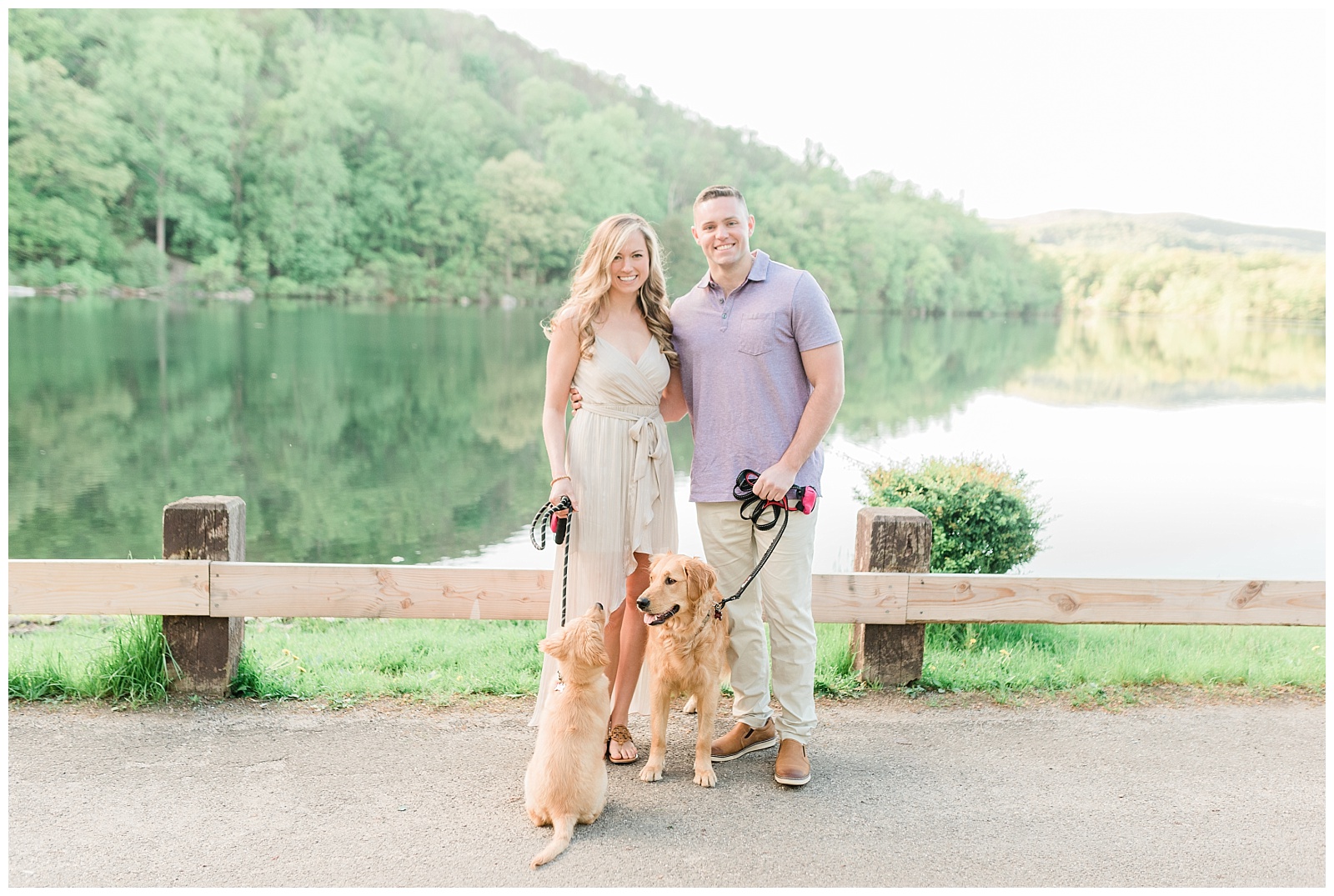 A couple stands with two golden retriever puppies on leashes in front of a lake.