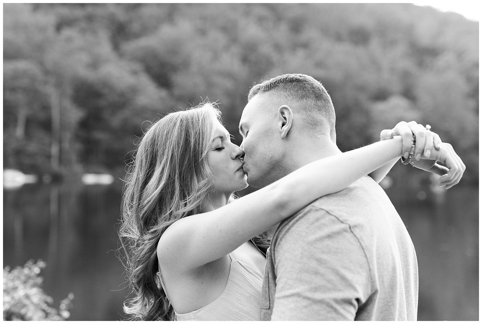 A couple kisses in front of a lake, in black and white.