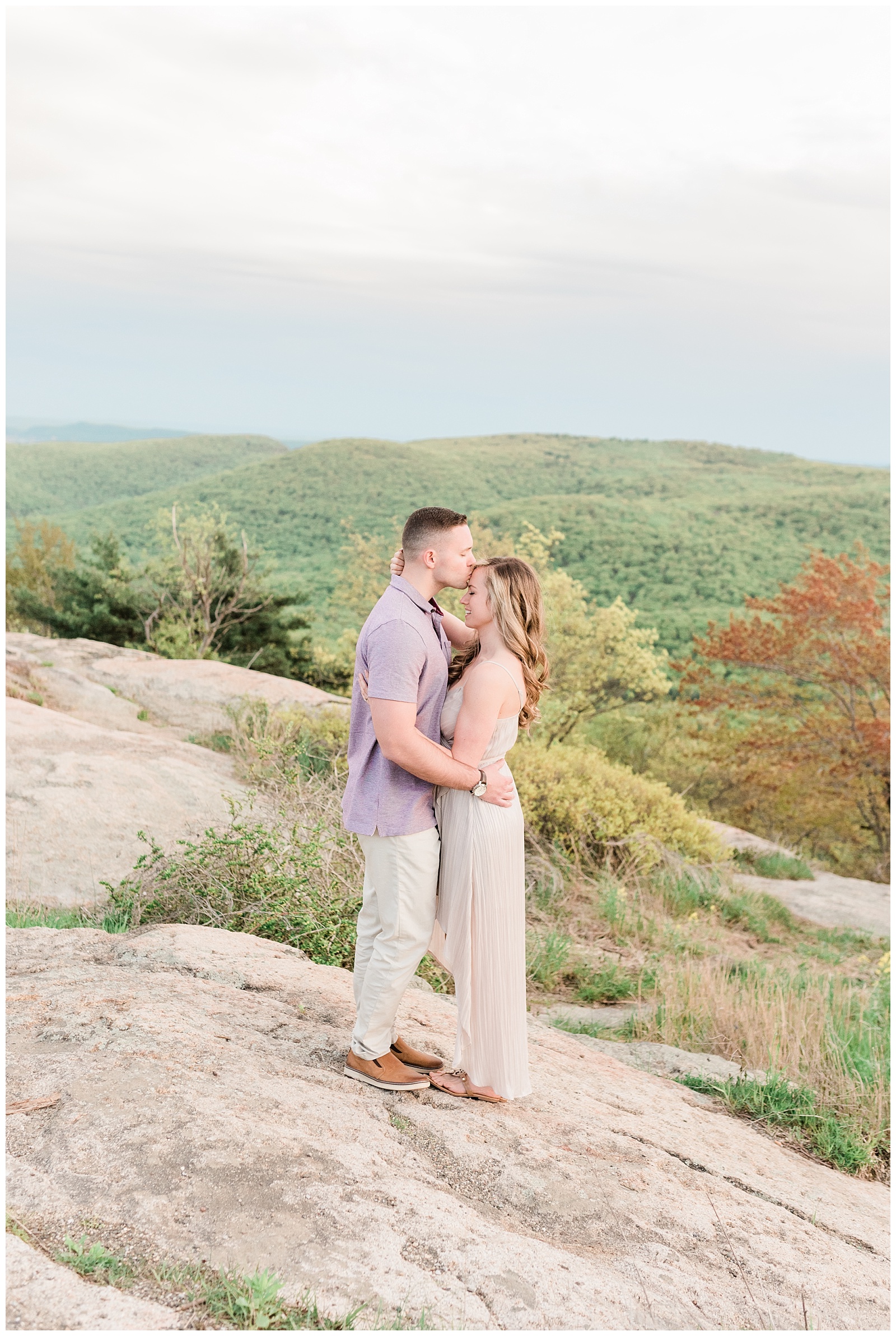 A man kisses a woman's forehead on top of a mountain during golden hour.