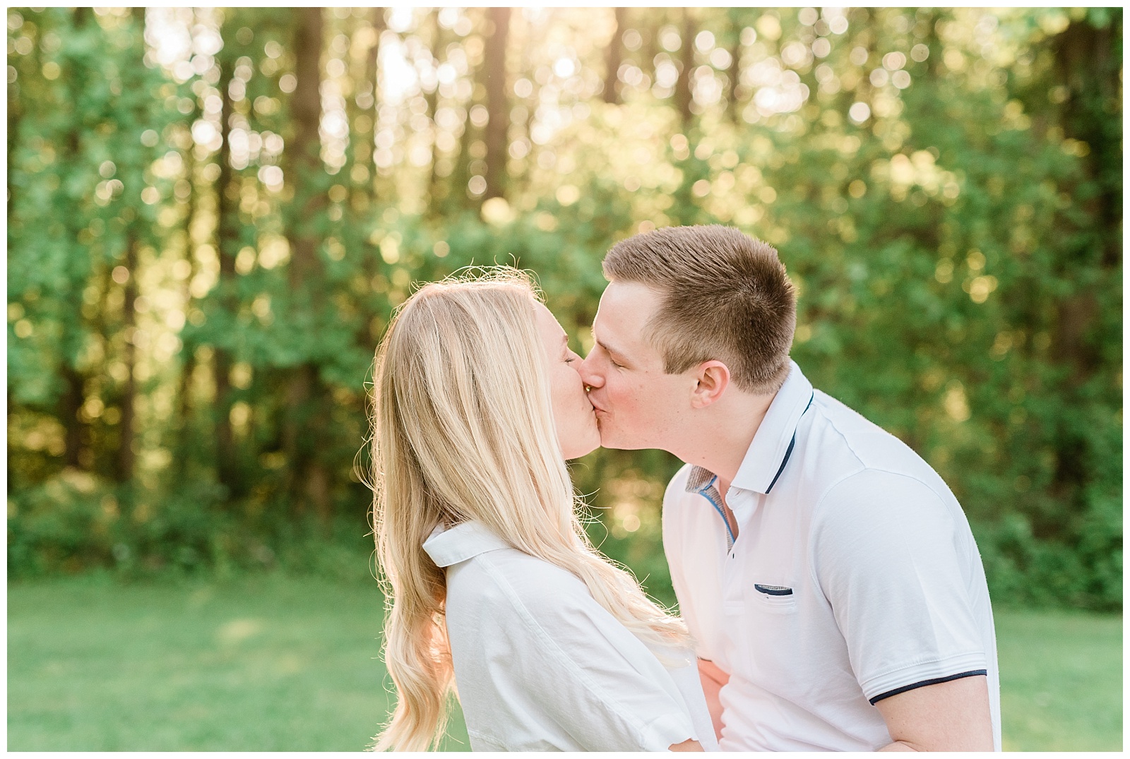 A couple kisses in front of a sunlit forest.