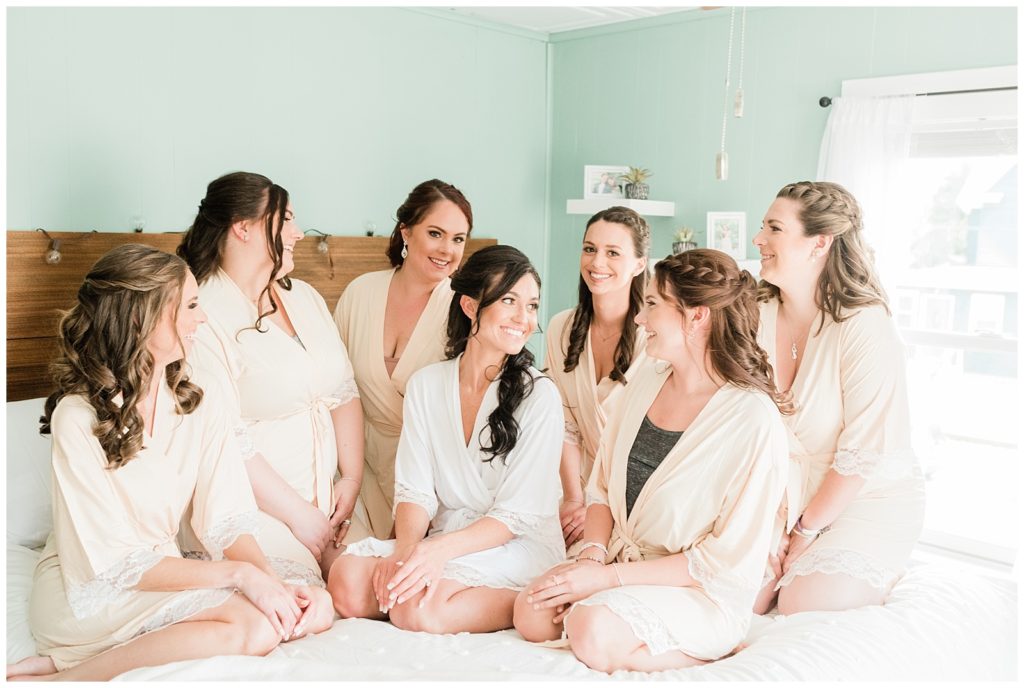 A bride and her bridesmaids wearing matching robes sit on the bed.