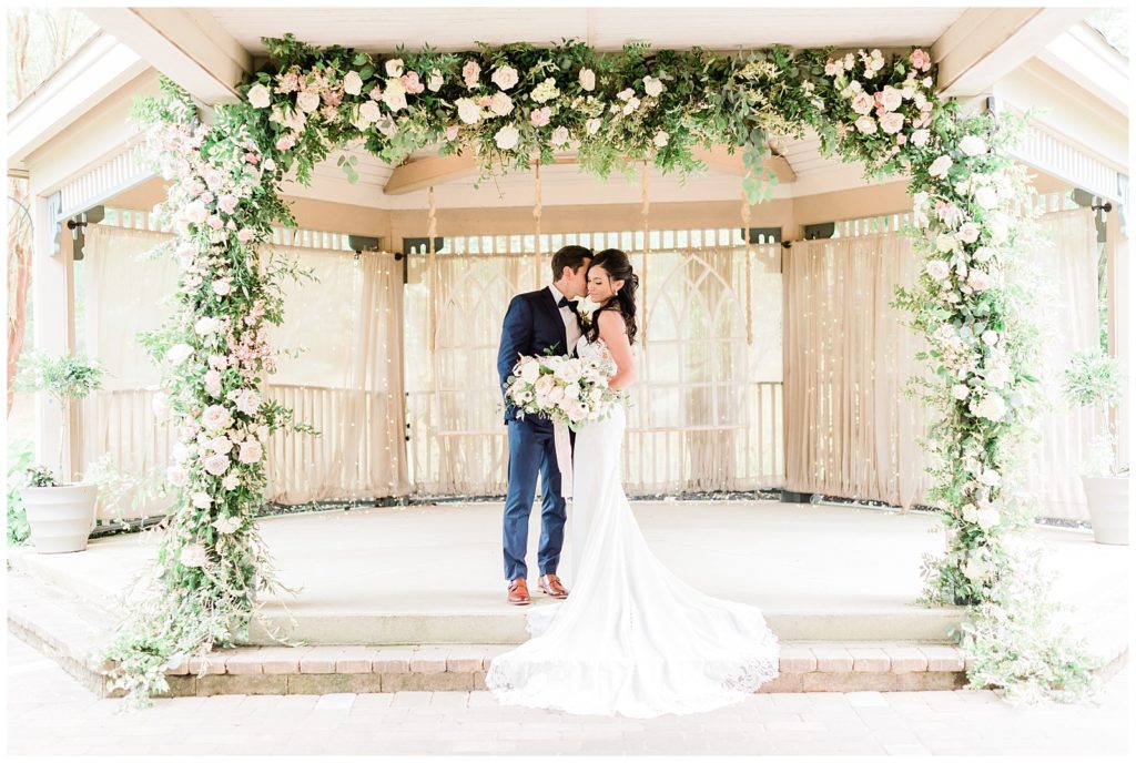 A groom nuzzles into the bride's temple while standing beneath a flower arch.