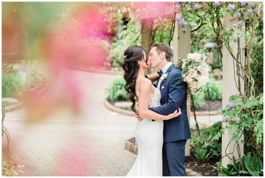 A bride and groom kiss beneath a flower covered pergola.