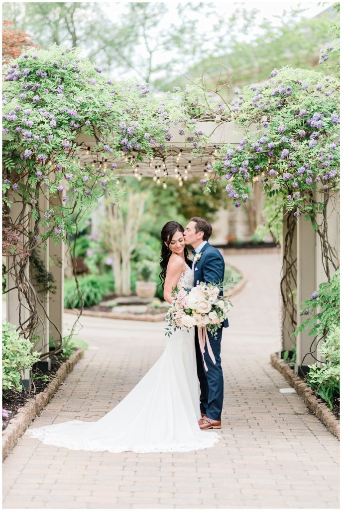 A groom kisses his bride on the cheek beneath a pergola covered in flowers.
