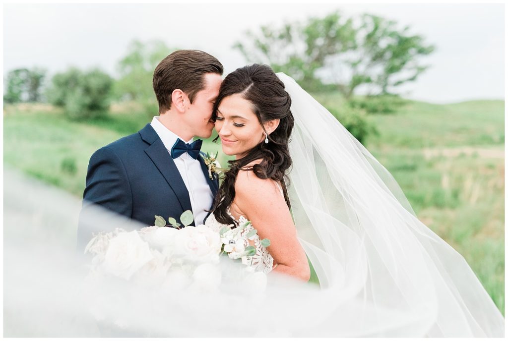 The groom kisses the bride on her cheek while her veil flies in the wind.