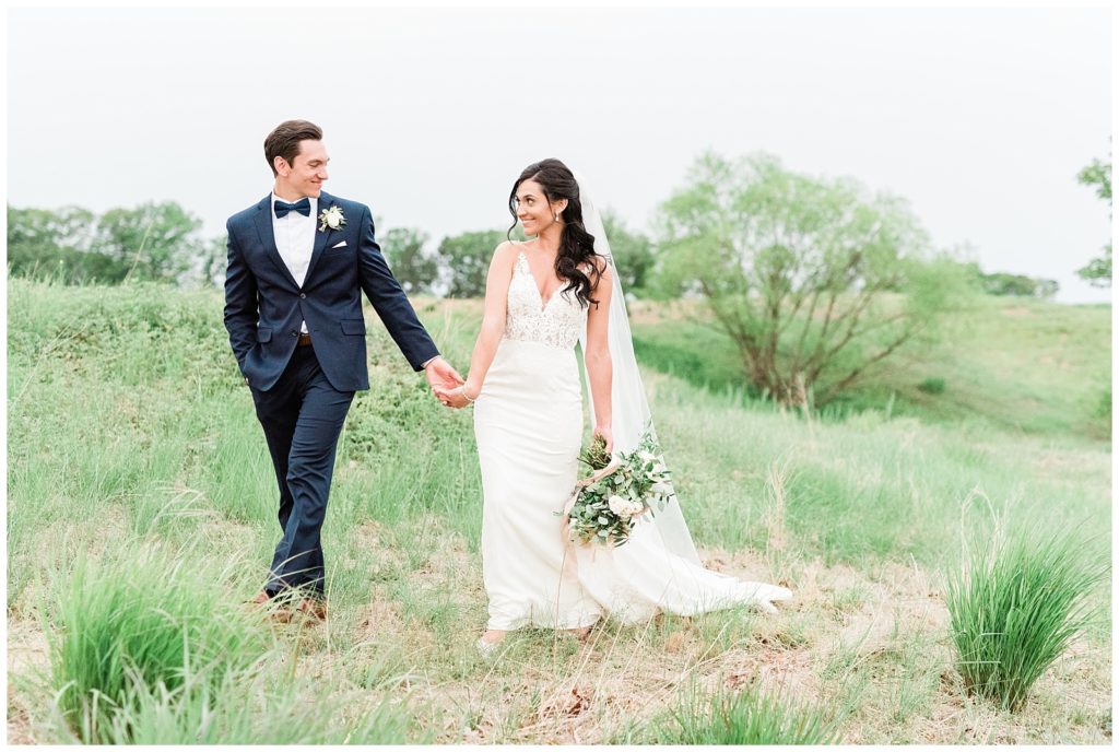 The groom holds his bride's hand and leads her through a grassy field.