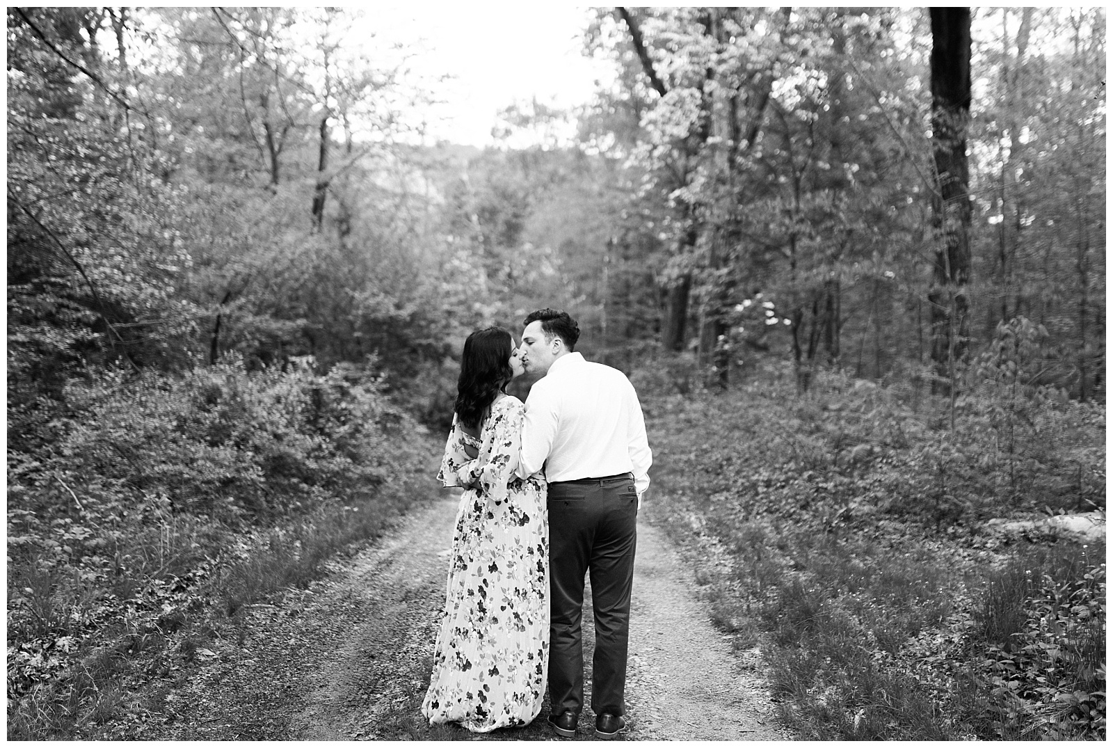A couple stands on a path in the woods kissing.