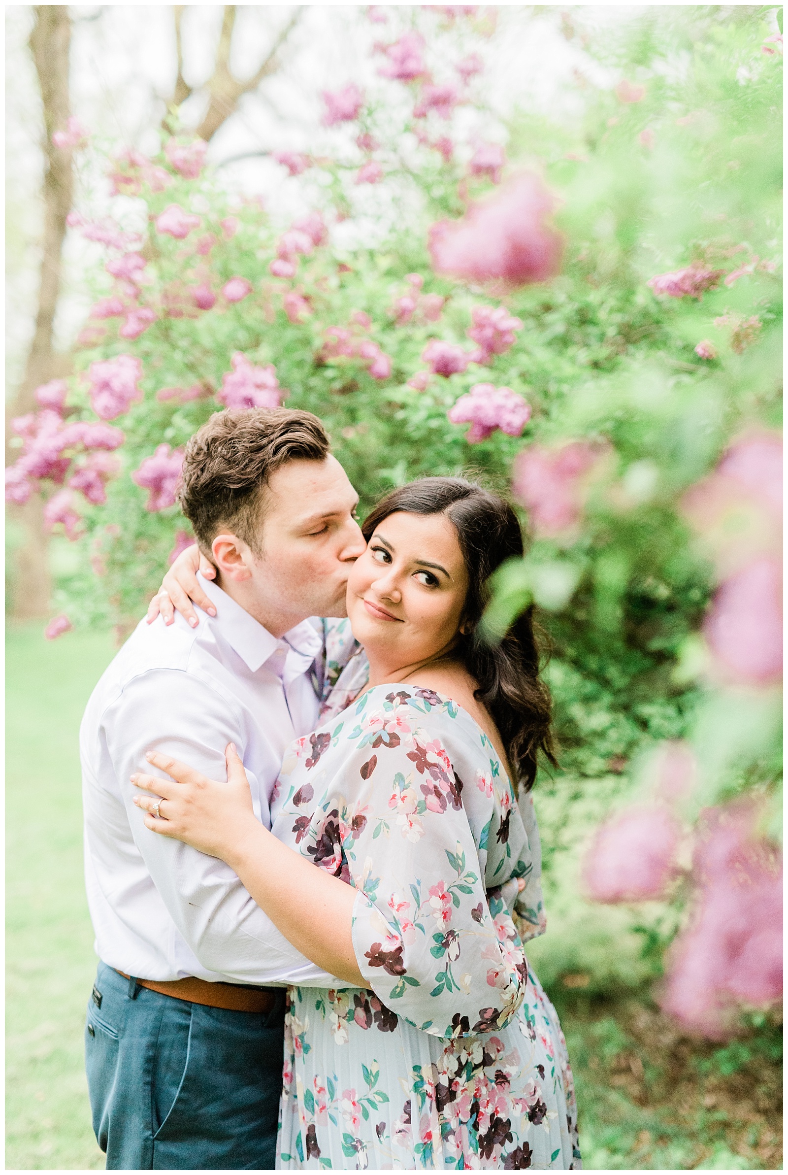 A man kisses a woman's cheek in the middle of lilac trees in a botanical garden.