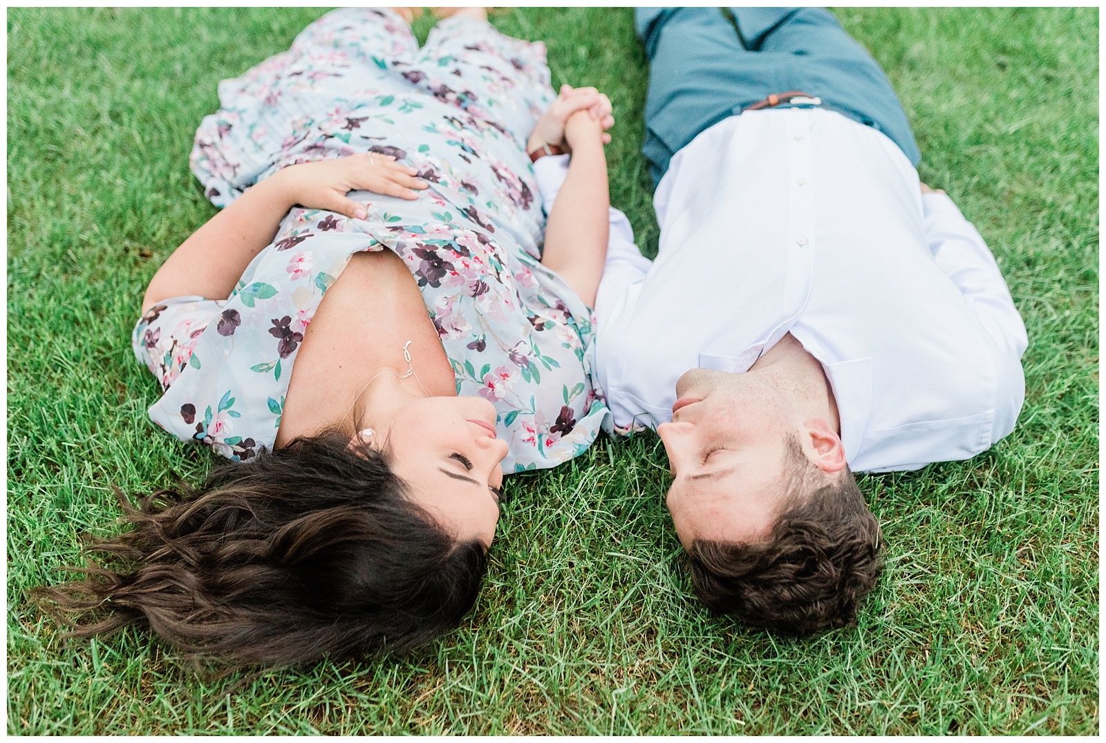 A couple holds hands laying in the grass while looking at each other.