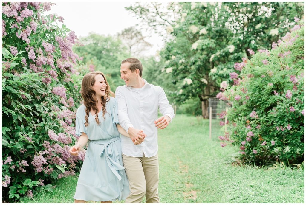 A couple walks through a lilac field holding hands and laughing at Willowwood Arboretum in NJ.