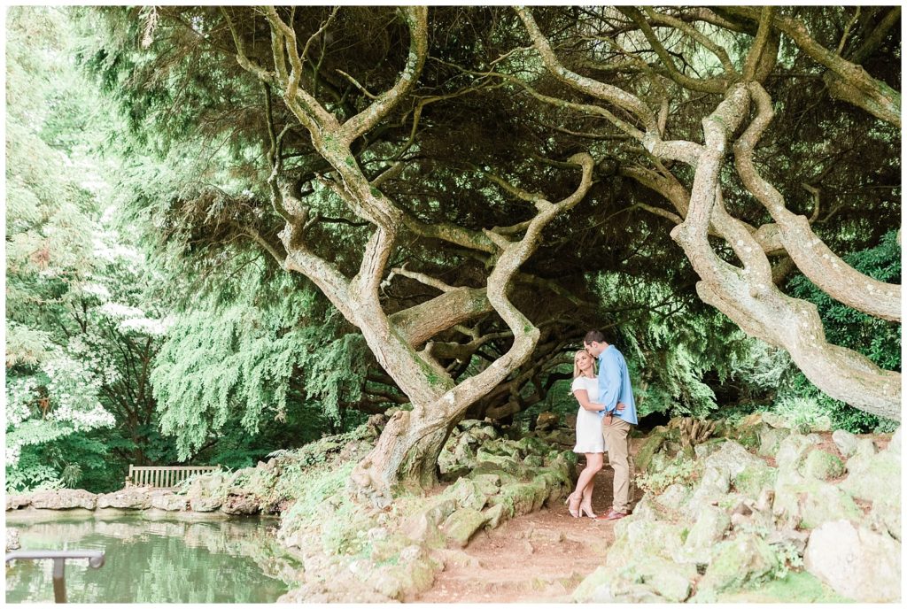 A man kisses his fiances cheek beneath large twisted trees in Deep Cut Gardens in NJ.