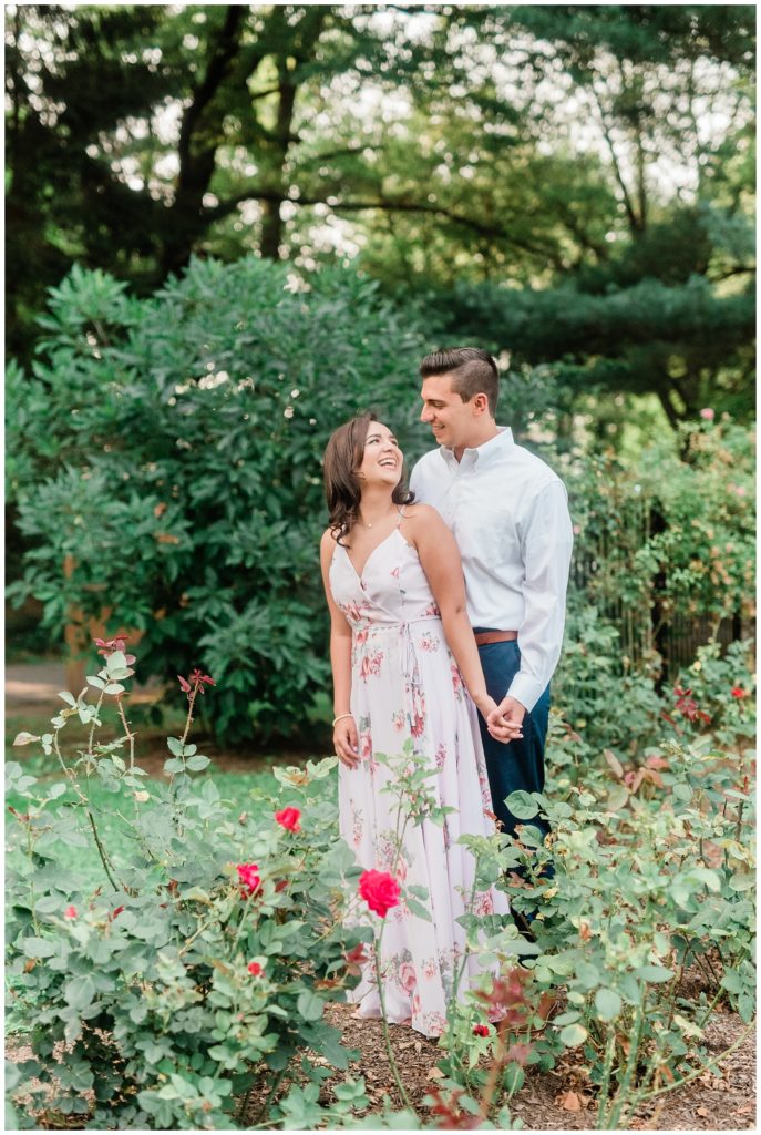 A couple holds hands standing among blooming roses in the garden.