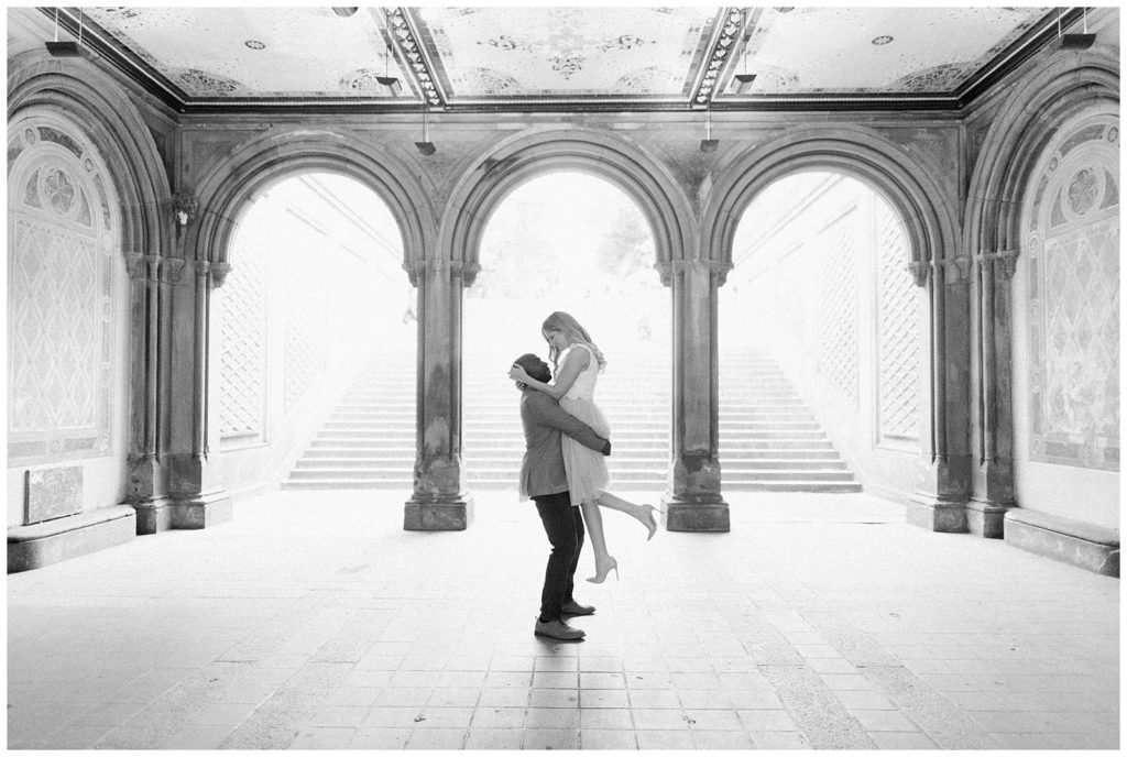 A man lifts his fiance up inside the Bethesda Terrace with the three archways in the background.