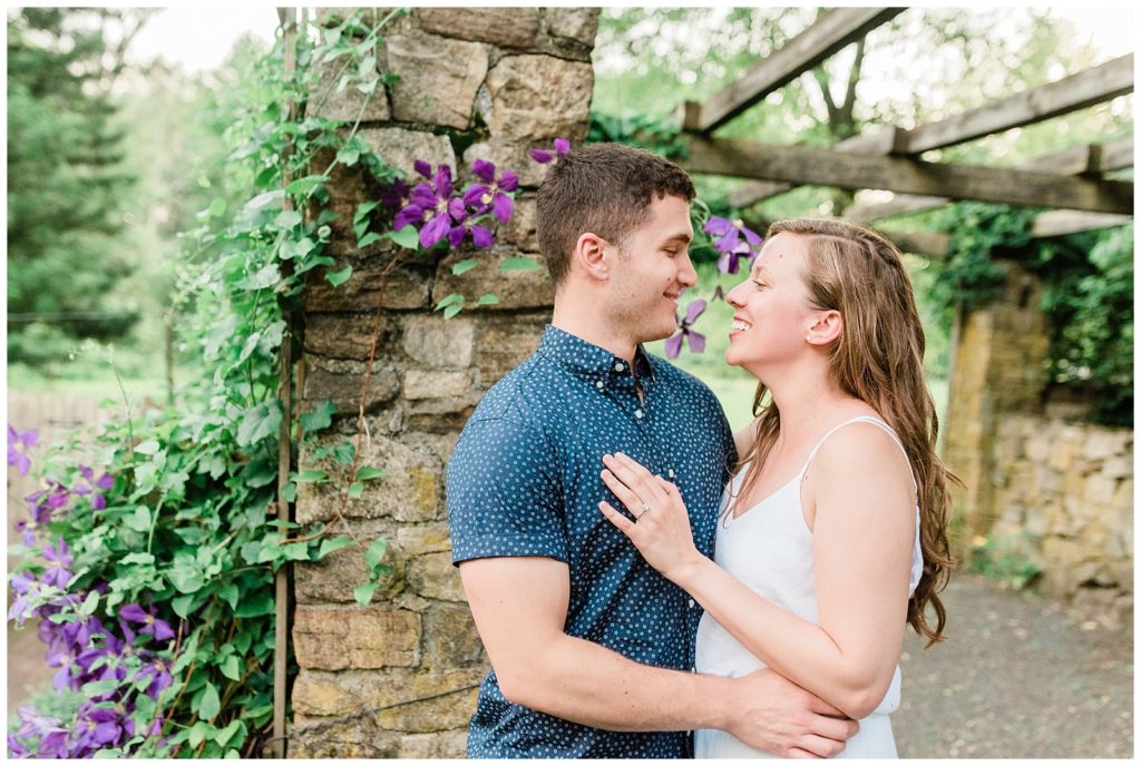 A couple looks at eachother with vines of purple flowers climbing the wall behind them.