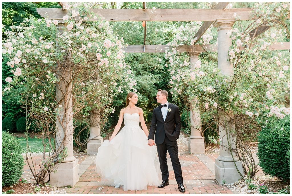 A bride and groom hold hands walking through a rose garden.