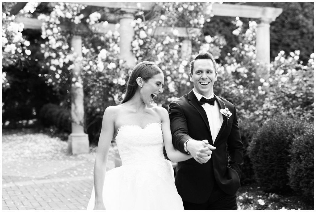 A bride and groom hold hands walking and laughing through a rose garden.