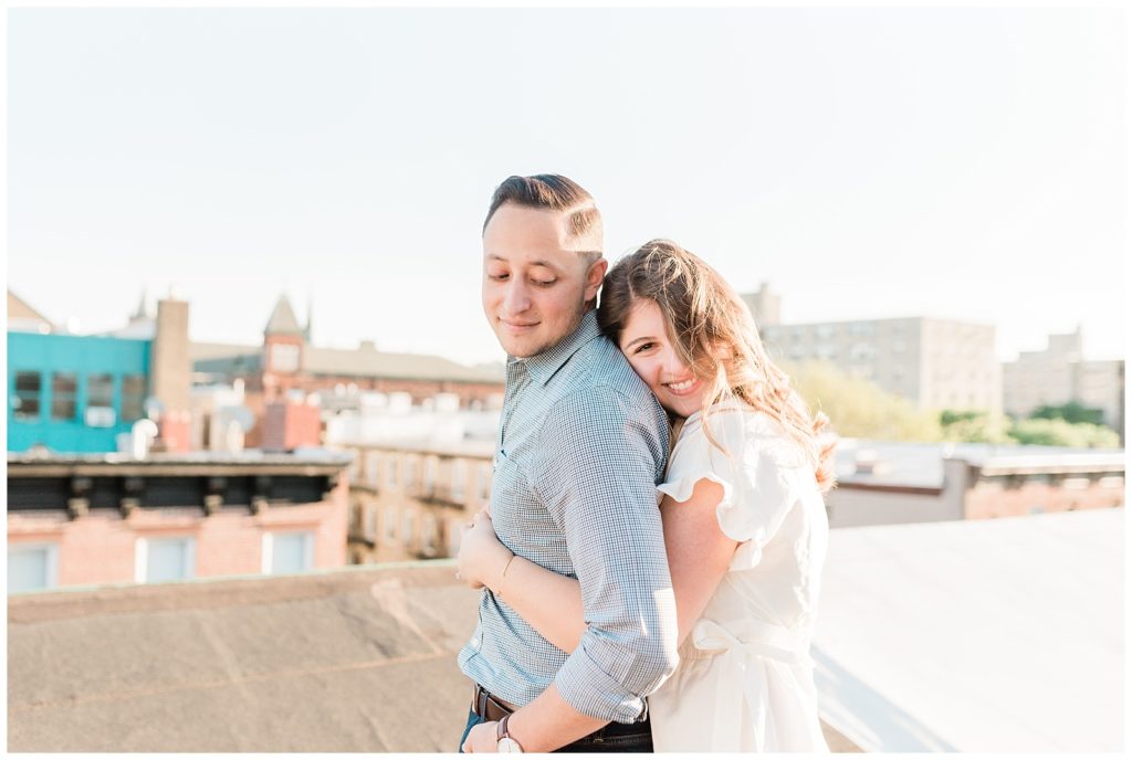 A woman hugs her fiance from behind on a rooftop in Hoboken, NJ